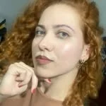 Michelle Fortes, ruiva de cabelo cacheado, com a mão perto do rosto
